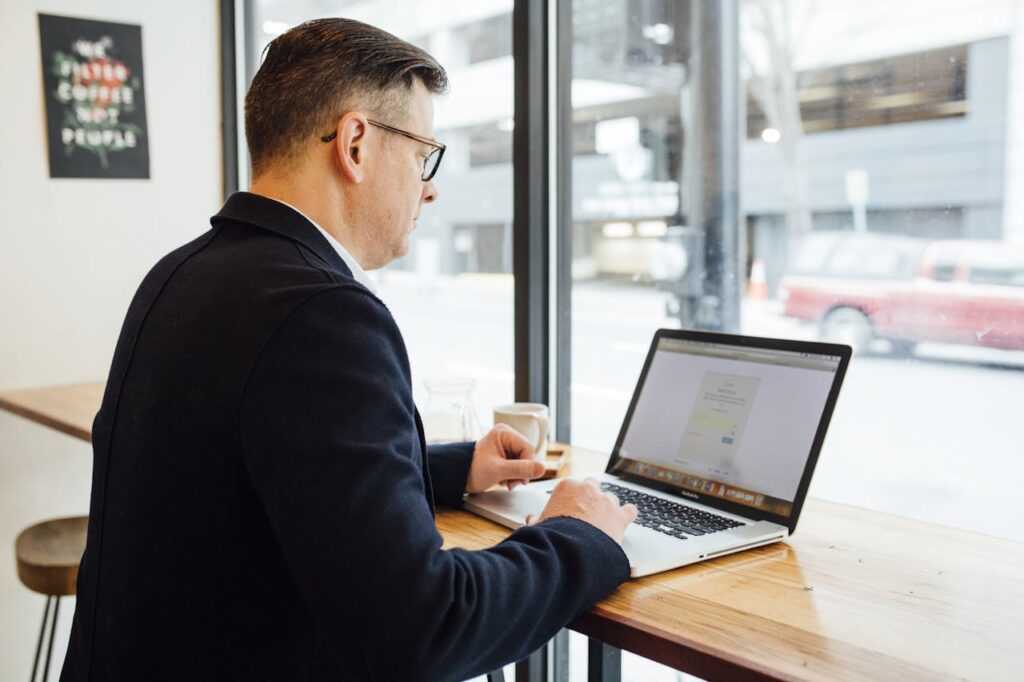 Adult male in cafe working on laptop, drinking coffee. Bright workspace with street view.
